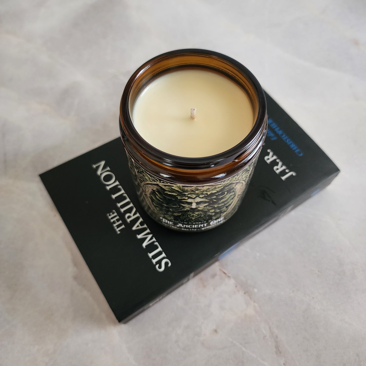 Ancient One soy fantasy candle marble surface placed on a book called the Silmarillion, handcrafted by Viridian Candle Co. Fantasy themed candle inspired by mythology.