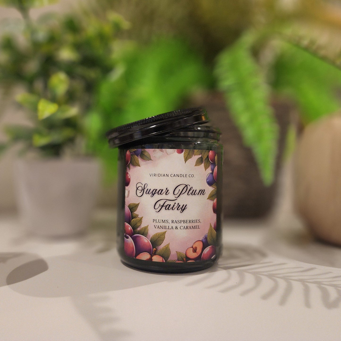 Sugar Plum Fairy soy fantasy candle with notes of plums and honey on a white surface with plants in the background. fantasy inspired candle by Viridian Candle Co.