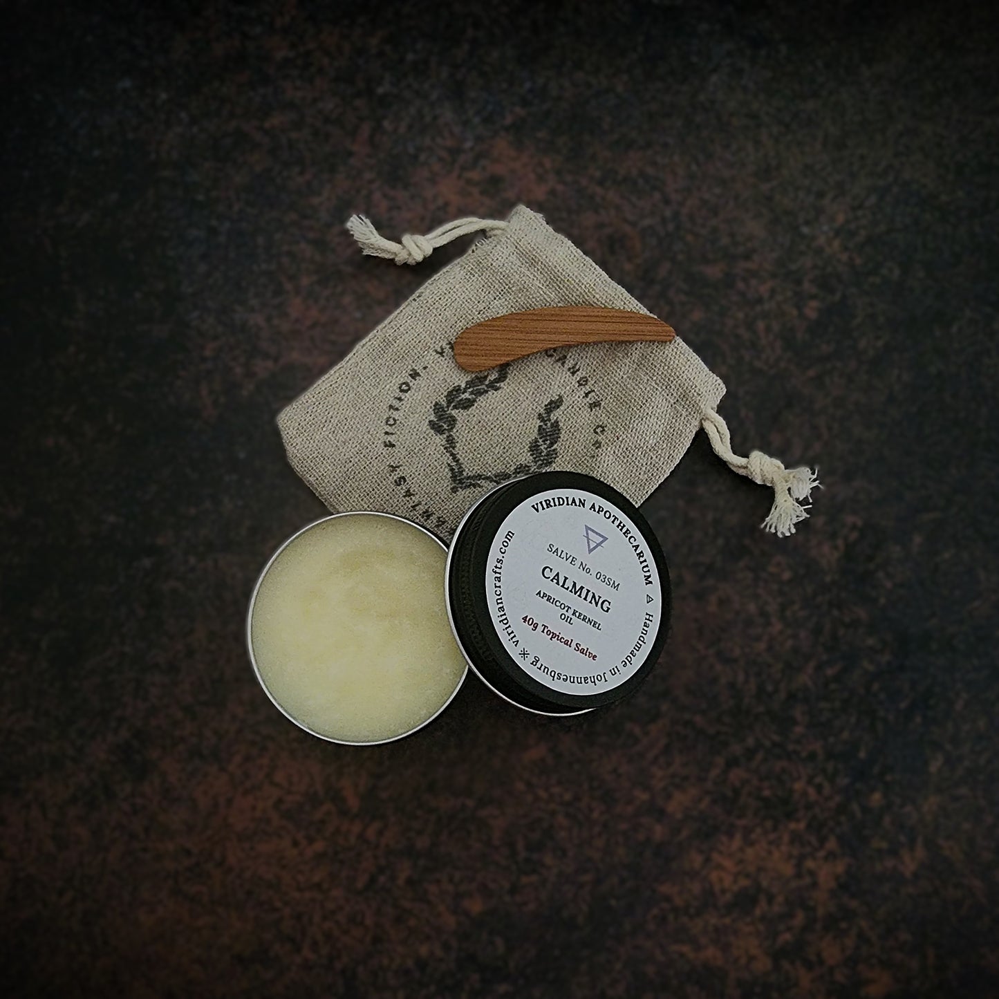 An opened tin of Calming Salve by Viridian Apothecarium, with a drawstring bag and bamboo spatula on a textured dark surface.