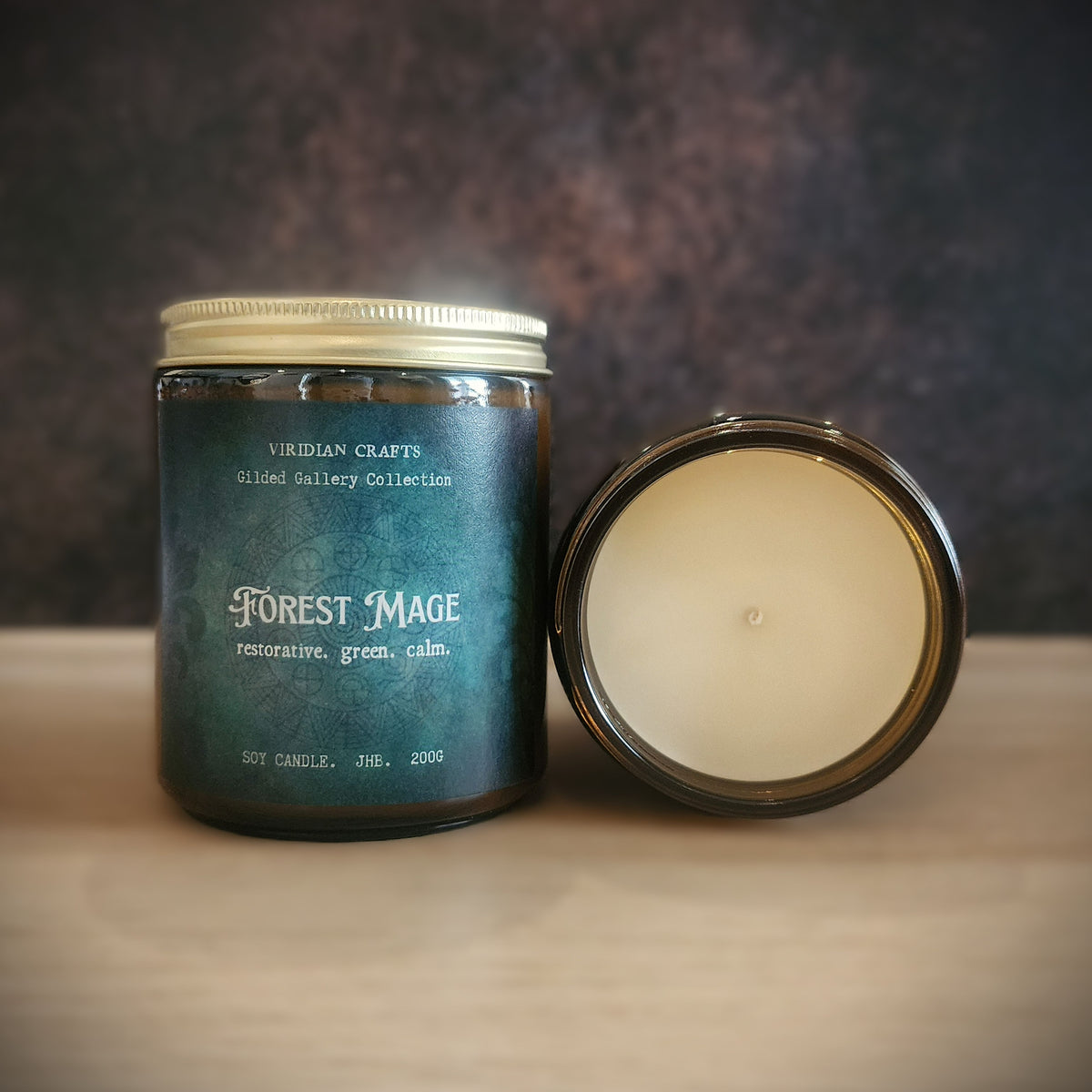 A jar candle labeled 'Forest Mage' by Viridian Crafts, shown with the dark teal glass jar and its cream-colored wax lid placed beside it on a light wooden surface against a dark textured background.