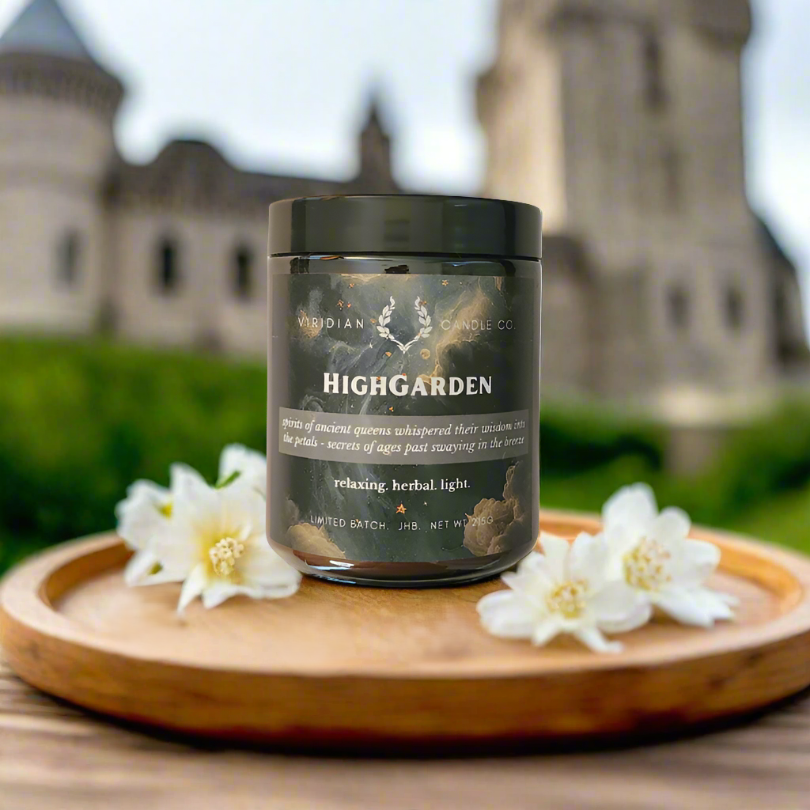 A HighGarden soy candle inspired by GoT's House Tyrell, and handcrafted by Viridian Candle Co., placed on a wooden tray in the garden with a castle in the background.