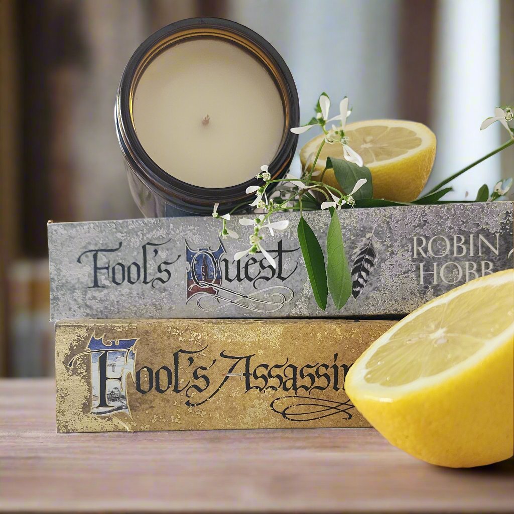 Royal Fool soy fantasy candle inspired by epic fantasy books, handcrafted by Viridian Candle Co. on a pile of Robin Hobb books with freshly cut lemons. Bright fruity nostalgic scent.