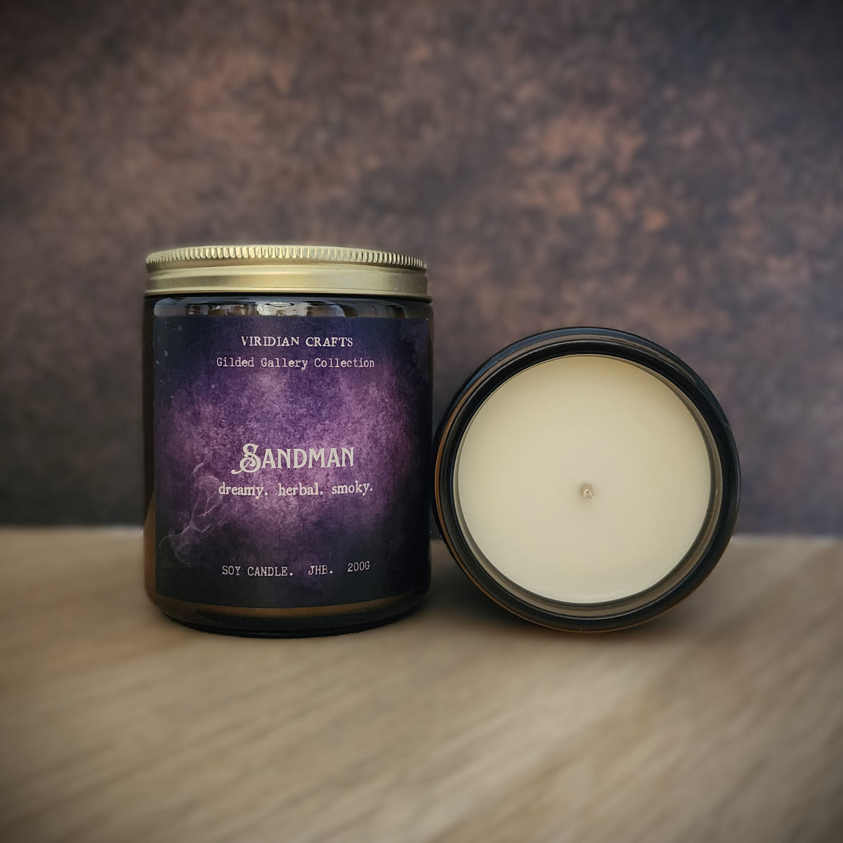 A scented candle in a dark glass jar with a gold lid, shown next to its open lid revealing cream-colored wax. The label features a purple galaxy design with the brand name 'VIRIDIAN CRAFTS' and product name 'SANDMAN'.