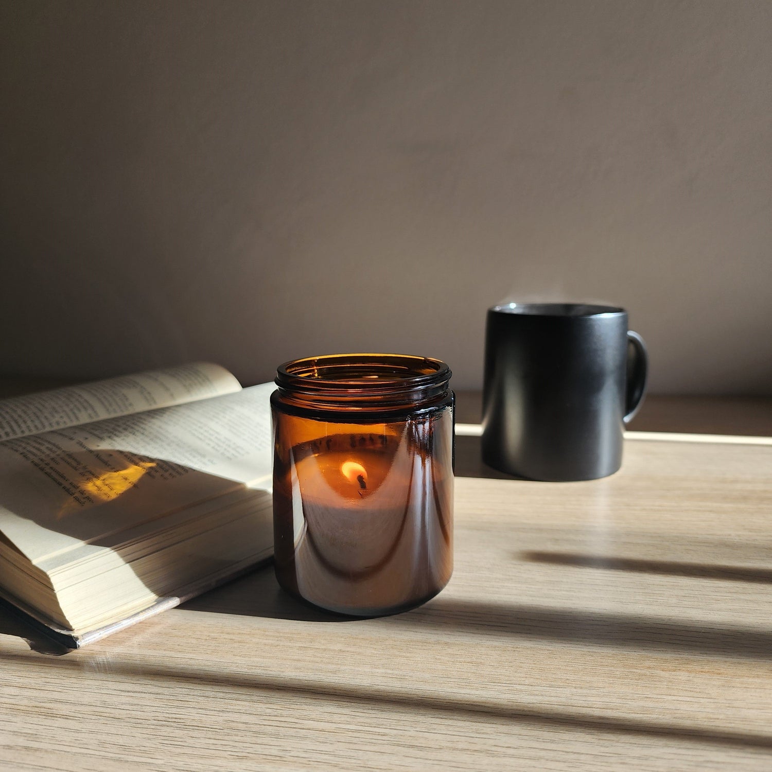 Soy candle on a table casting a shadow on an open book with a black steaming coffee mug in the background