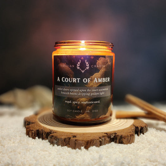 A soy candle labeled 'A Court of Amber' by Viridian Crafts, lit and placed on a sliced wood coaster with a dark blurred background and a faux fur surface.
