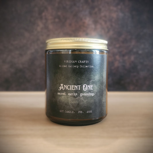 A soy candle in a dark amber glass jar with a gold metal lid, labeled 'Ancient One' by Viridian Crafts, set on a light wooden surface against a dark textured background.