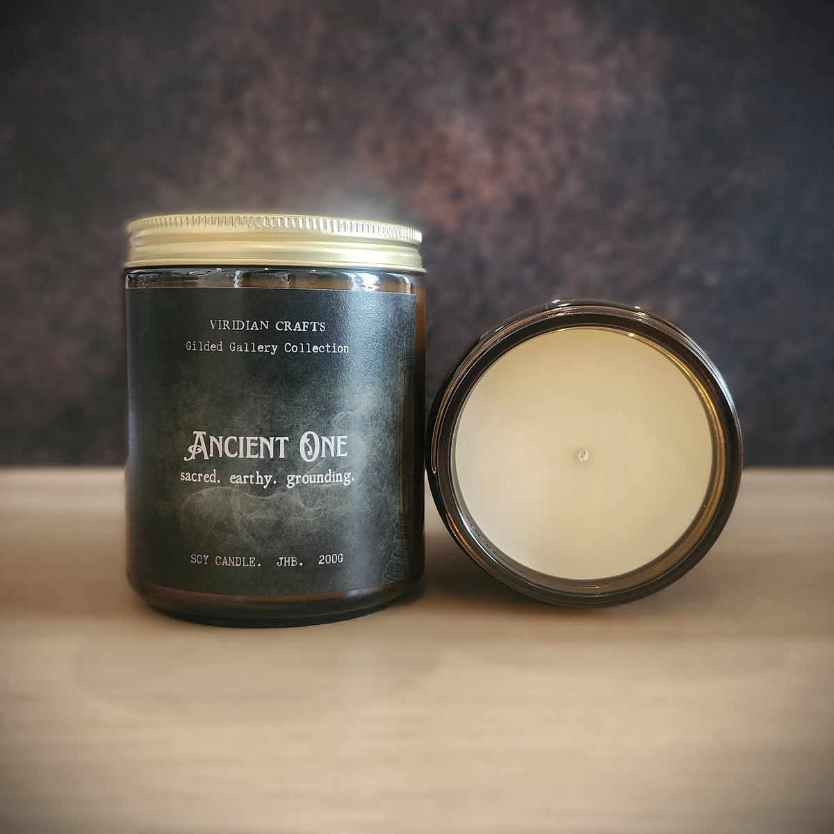 A dark amber glass jar candle labeled 'Ancient One' by Viridian Crafts, shown with its cream-colored wax visible in the open lid next to the closed jar on a wooden surface.