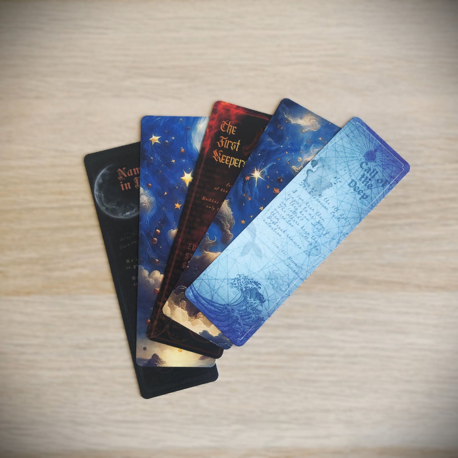 Four fantasy-themed bookmark cards fanned out on a light wood surface. The bookmarks feature celestial and ocean illustrations, including designs for Call of the Deep, The First Keepers and Names in Red
