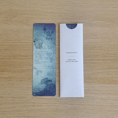 A rectangular blue and white paper bookmark with a wave illustration and the text 'Call of the Deep' is shown next to its white envelope-style packaging on a light wood surface.