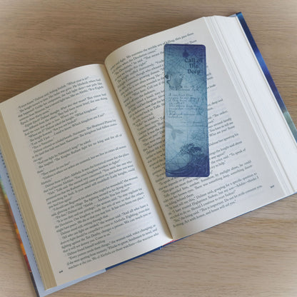 A decorative blue bookmark with a vintage map design and the text 'Call of the Deep' is placed on an open book with printed pages, photographed on a light wood surface.