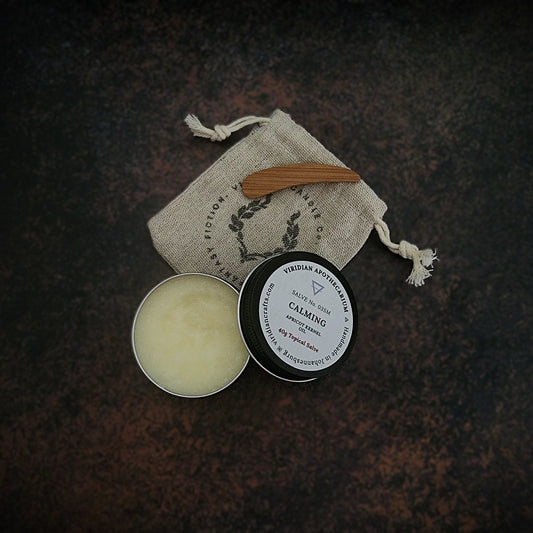 An opened tin of Calming Salve by Viridian Apothecarium, with a drawstring bag and bamboo spatula on a textured dark surface.