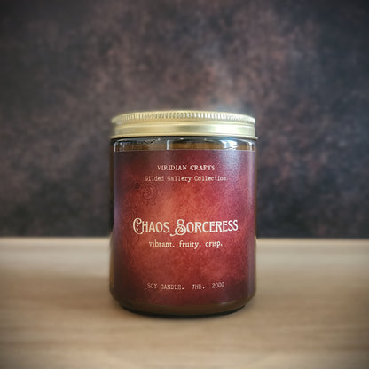 A glass jar candle with a metallic gold lid and a dark red label reading 'Chaos Sorceress' by Viridian Crafts, set on a light wooden surface against a dark textured background.