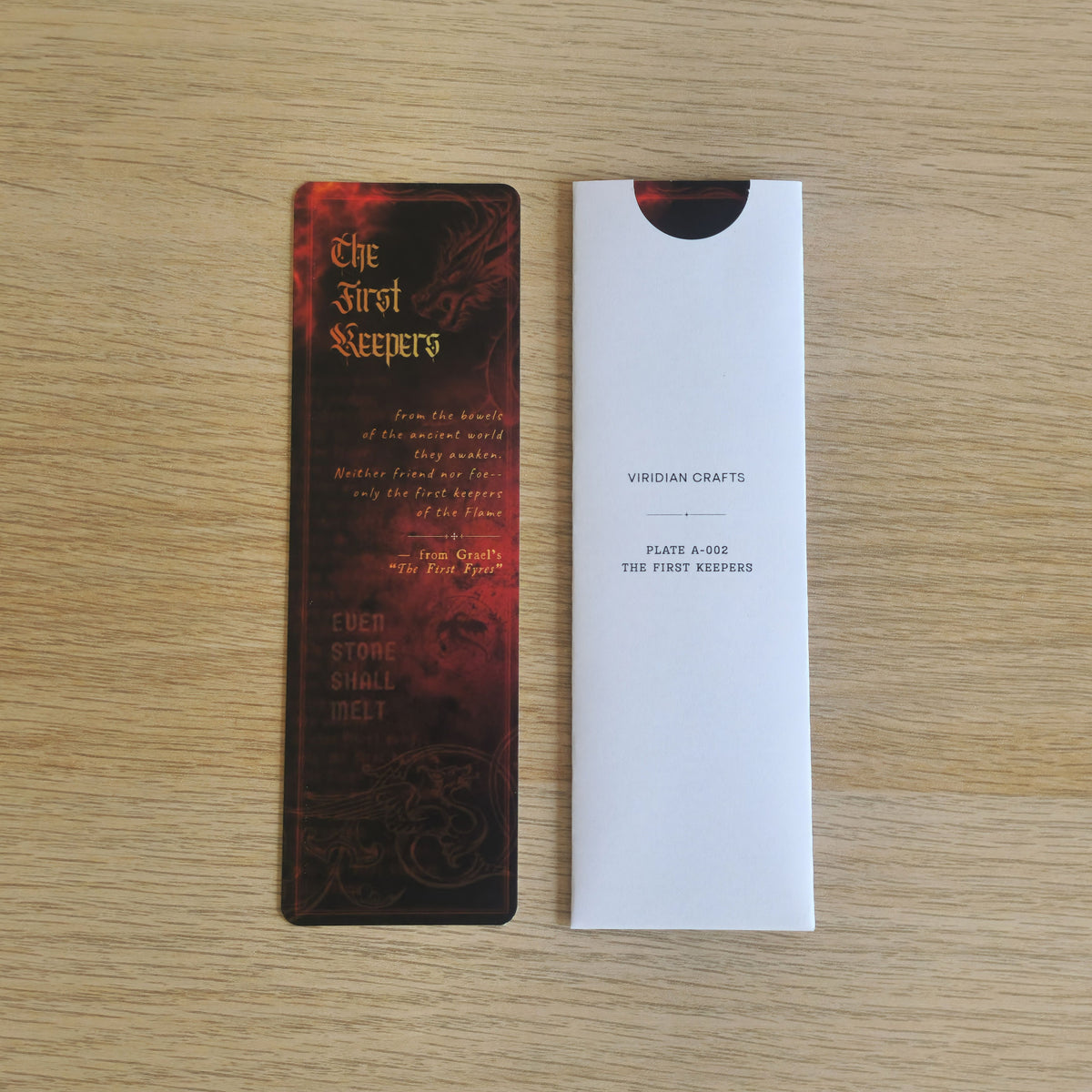 A rectangular bookmark with a dark red and black fantasy-style design titled "The First Keepers" is shown next to a bookmark enclosed in a protective white sleeve on a light wood surface. The sleeve has small printed text including "VIRIDIAN CRAFTS" and "PLATE A-002 THE FIRST KEEPERS".