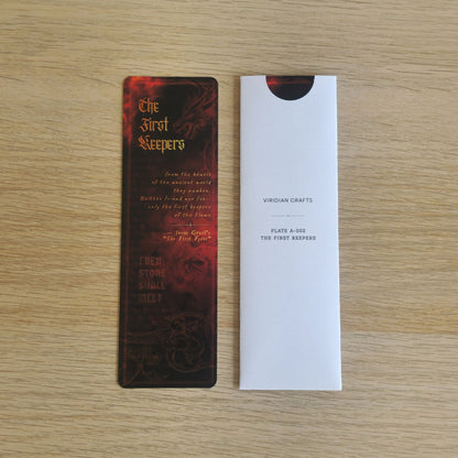 A rectangular bookmark with a dark red and black fantasy-style design titled "The First Keepers" is shown next to a bookmark enclosed in a protective white sleeve on a light wood surface. The sleeve has small printed text including "VIRIDIAN CRAFTS" and "PLATE A-002 THE FIRST KEEPERS".