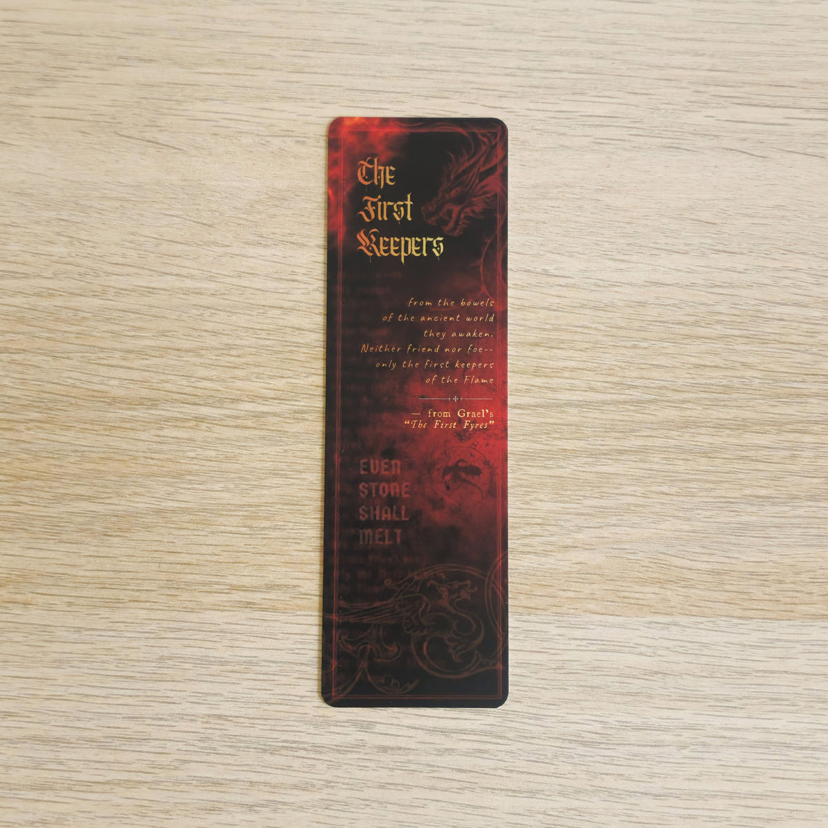 A slim rectangular book bookmark with a dark red and black dragon design and gold text reading 'The First Sleepers' and 'Even Stone Shall Melt,' placed on a light wood surface.