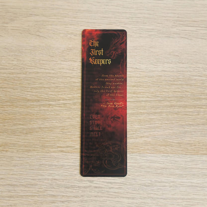 A slim rectangular book bookmark with a dark red and black dragon design and gold text reading 'The First Sleepers' and 'Even Stone Shall Melt,' placed on a light wood surface.
