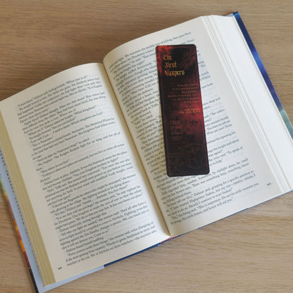 A dark red and black bookmark with gold lettering rests on the pages of an open book on a light wood surface.