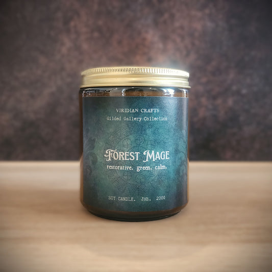 A soy candle in a clear glass jar with a gold metal lid, featuring a dark teal label with white text that reads 'Forest Mage' and 'restorative, green, calm.' by Viridian Crafts, set on a light wooden surface against a dark textured background.