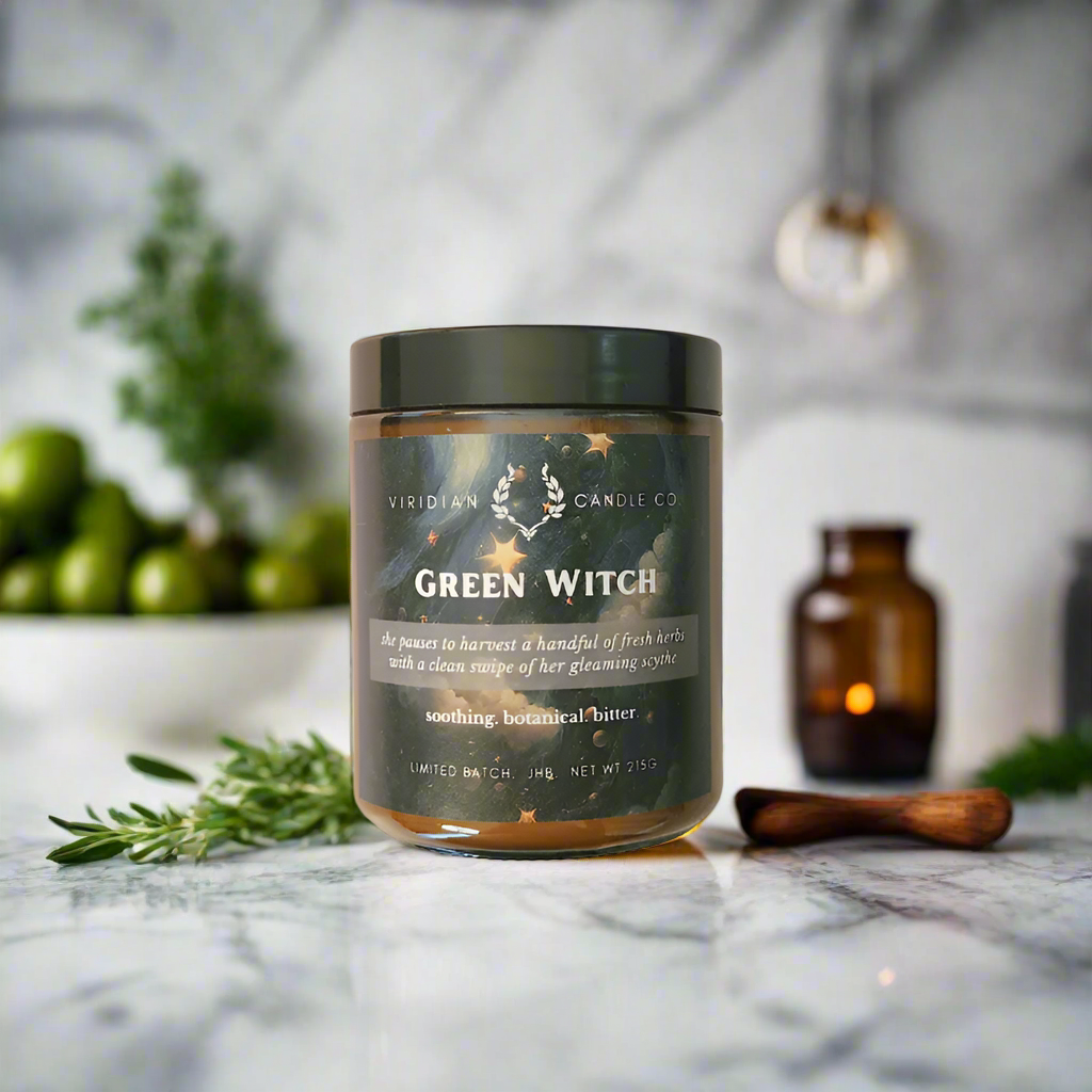 A Green Witch soy fantasy candle by Viridian Candle Co., placed on a marble surface with a decorative item and a wooden spoon in the background. Fantasy themed candle