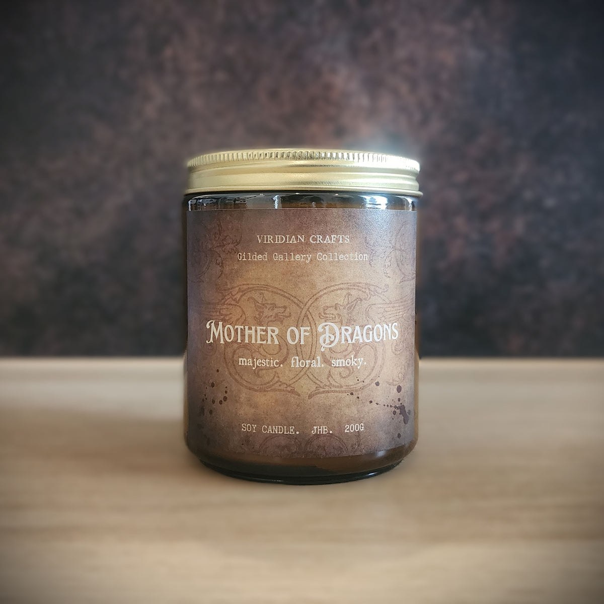 A soy candle in a dark glass jar with a metallic gold lid, labeled 'Mother of Dragons' by Viridian Crafts, set on a light wooden surface against a dark textured background.