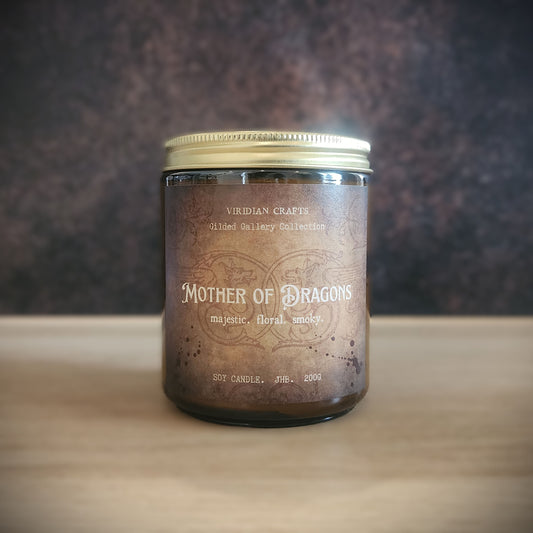 A soy candle in a dark glass jar with a metallic gold lid, labeled 'Mother of Dragons' by Viridian Crafts, set on a light wooden surface against a dark textured background.