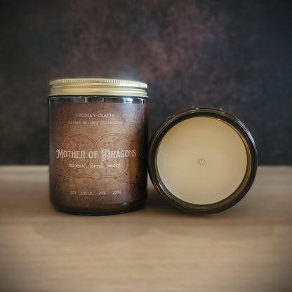 A jar candle labeled 'Mother of Dragons' by Vidian Crafts sits on a light wood surface with its lid placed beside it. The candle jar is dark amber-brown glass with a cream-colored wax insert visible inside the open lid, and a metallic gold-toned lid sits on top of the closed jar.
