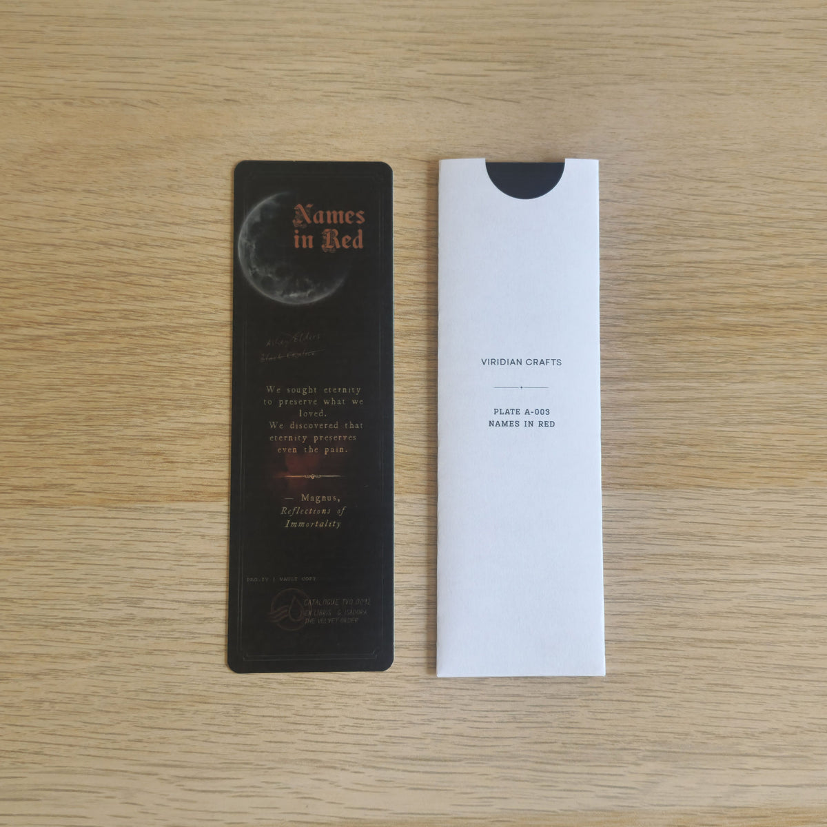 A black bookmark with the title 'Names in Red' and a moon graphic, placed next to a bookmark enclosed in a protective white sleeve with 'Viridian Crafts' printed on it, on a light wood background.