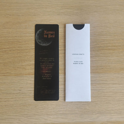 A black bookmark with the title 'Names in Red' and a moon graphic, placed next to a bookmark enclosed in a protective white sleeve with 'Viridian Crafts' printed on it, on a light wood background.
