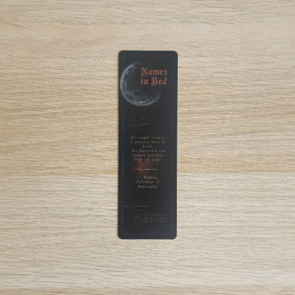 A slim black bookmark with a moon graphic and the title 'Names in Red' in orange-red text, placed on a light wood grain surface.