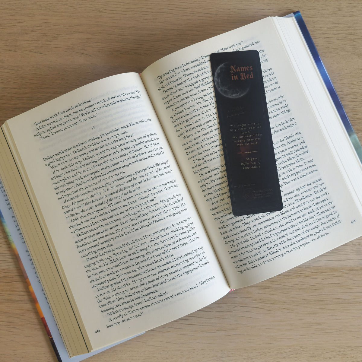 A black book bookmark with gold text reading 'Names in Red' and a crescent moon design, placed on an open book on a light wood surface.