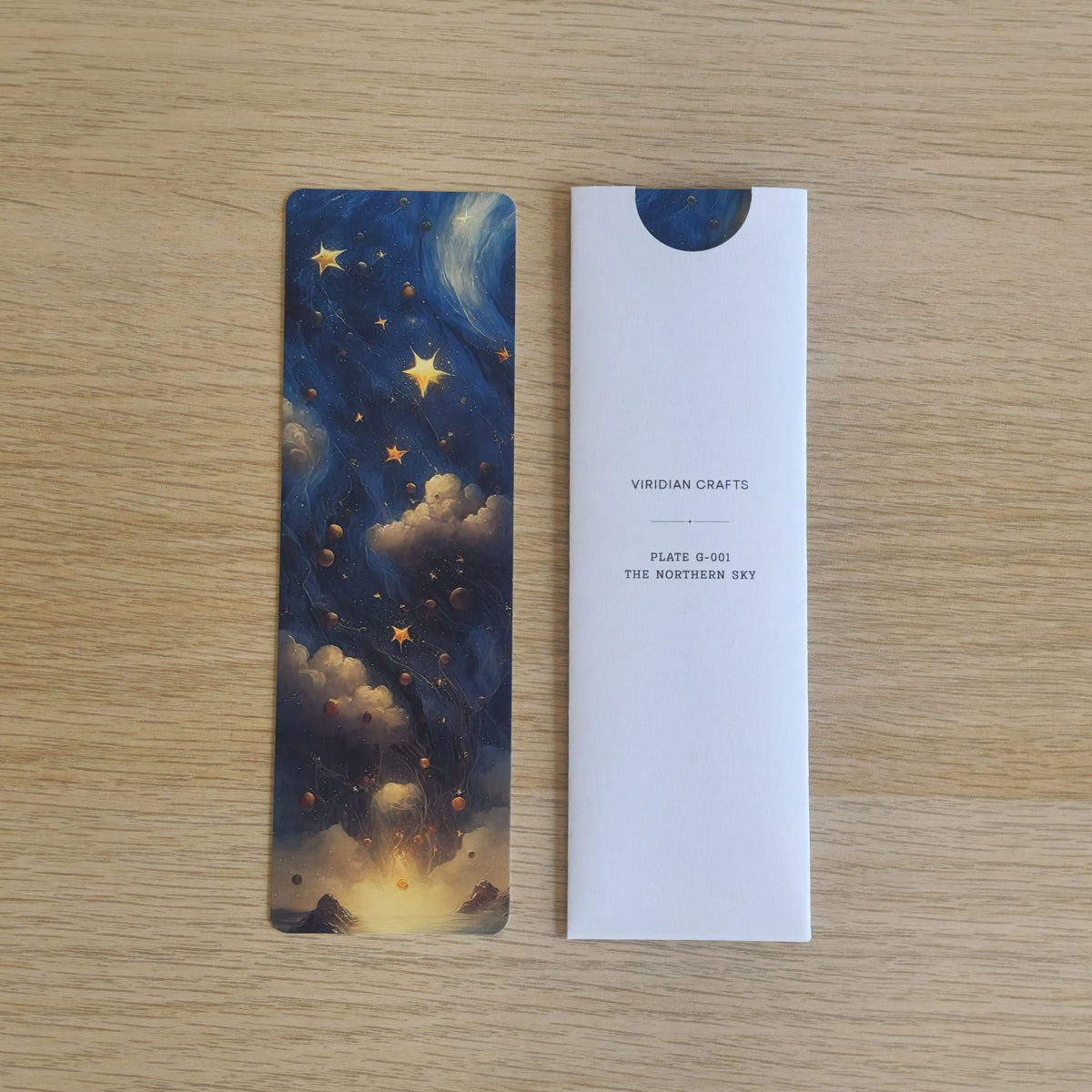 A rectangular bookmark with an illustrated night sky scene in blue, cream, and gold, placed beside a bookmark enclosed in a white protective paper sleeve printed with the brand name 'Viridian Crafts' and product text, on a light wood surface.