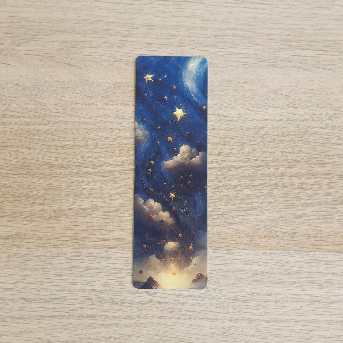 A slim bookmark with rounded corners, featuring an artistic night sky design with blue clouds, glowing yellow stars, and a warm light at the bottom, placed on a light wood surface.