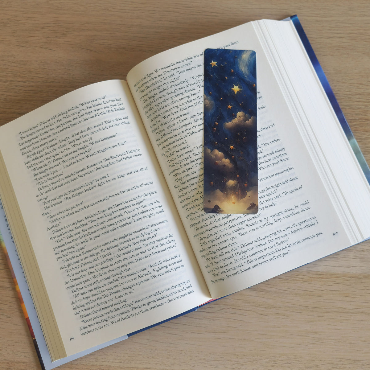 An open book on a light wood surface, with a decorative bookmark featuring a night sky and starry cloud design placed in the center of the right page.