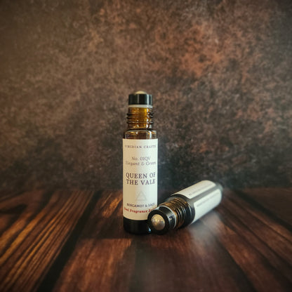 Queen of the Vale Essence - Perfume Roll-On 10ml amber bottle with a second bottle lying on its side containing a blend of bergamot and sage, handcrafted by Viridian Crafts on a moody industrial surface and background.