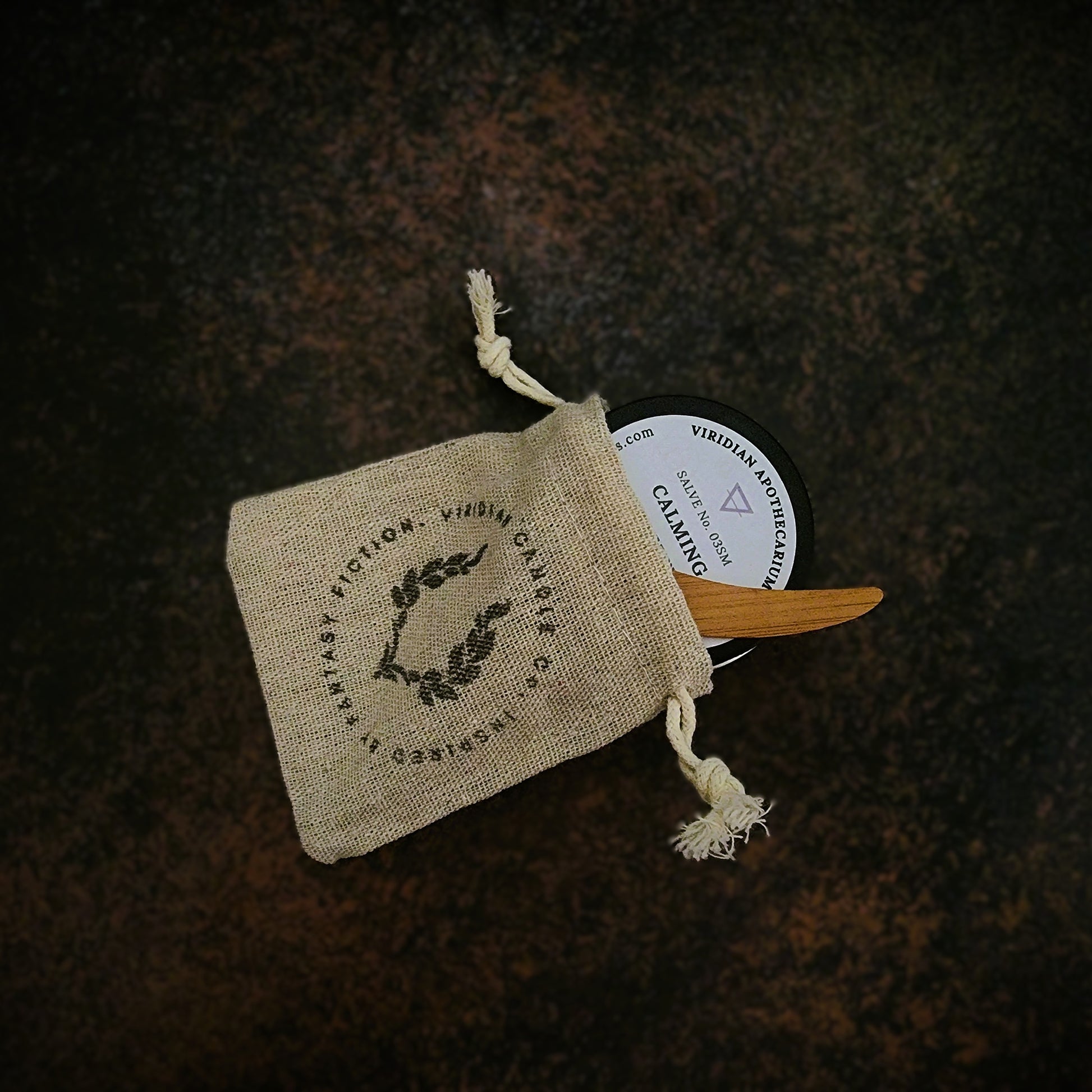 An tin of Calming Salve by Viridian Apothecarium, halfway in a burlap drawstring bag and bamboo spatula on a textured dark surface.