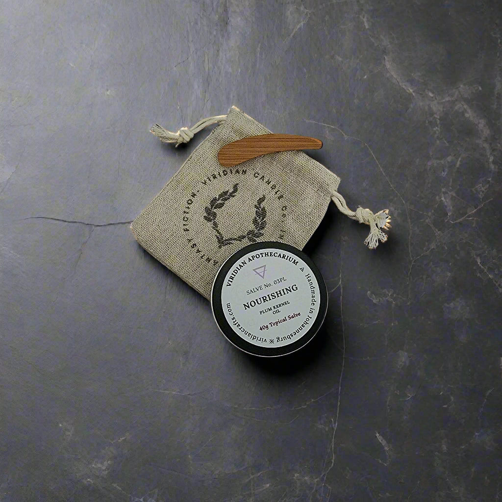 A tin of Nourishing Salve by Viridian Apothecarium, with a drawstring bag and bamboo spatula on a textured dark surface.