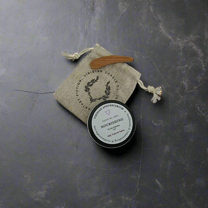 A tin of Nourishing Salve by Viridian Apothecarium, with a drawstring bag and bamboo spatula on a textured dark surface.