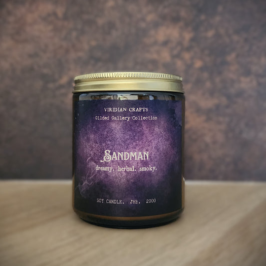 A jar candle with a purple watercolor-style label reading 'Sandman' and 'Viridian Crafts', set on a light wood surface against a textured brown background.