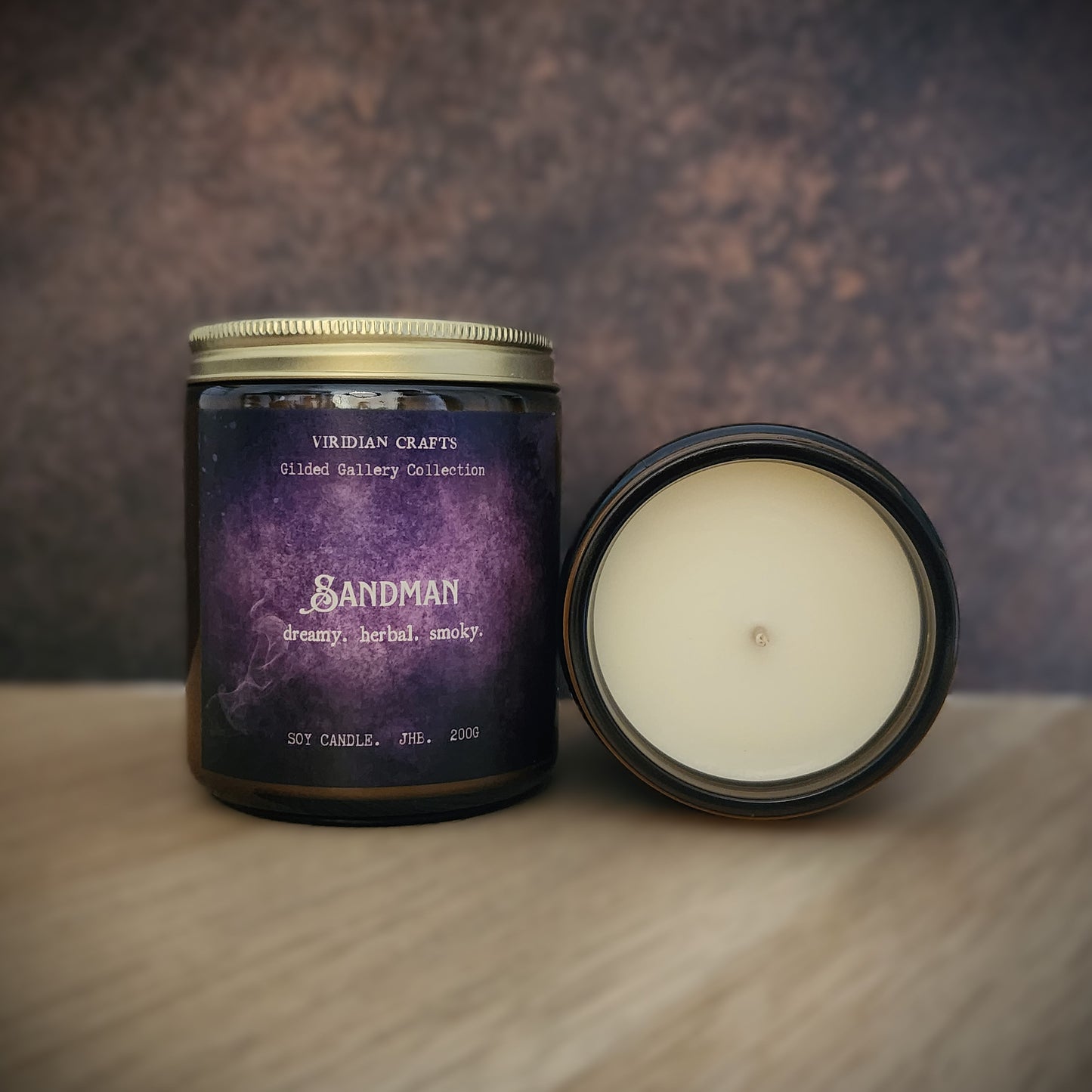 A scented candle in a dark glass jar with a gold lid, shown next to its open lid revealing cream-colored wax. The label features a purple galaxy design with the brand name 'VIRIDIAN CRAFTS' and product name 'SANDMAN'.