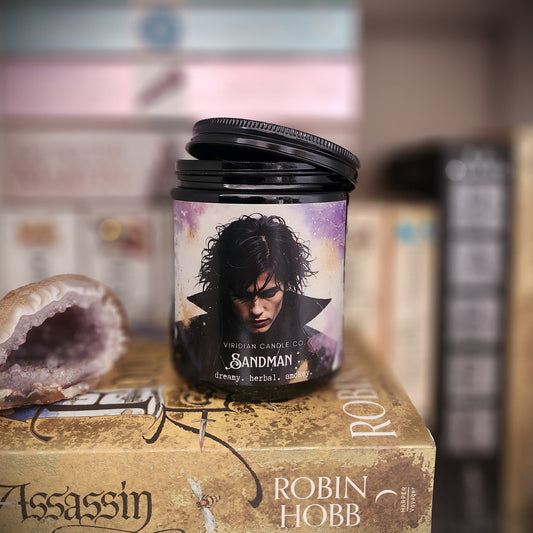 Sandman fantasy soy candle by Viridian Candle Co., on a bookcase with an amethyst geode next to it.