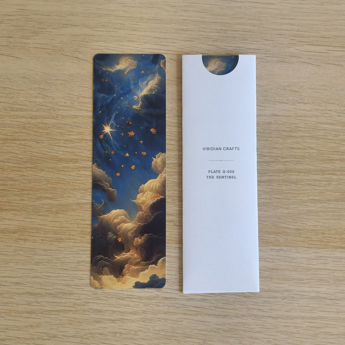 A rectangular bookmark with an illustrated night sky and clouds in dark blue, gold, and cream, placed beside a bookmark enclosed in a white protective paper sleeve printed with the brand name 'VIRIDIAN CRAFTS' and product text 'PLATE 8-602 THE SENTINEL' on a light wood surface.