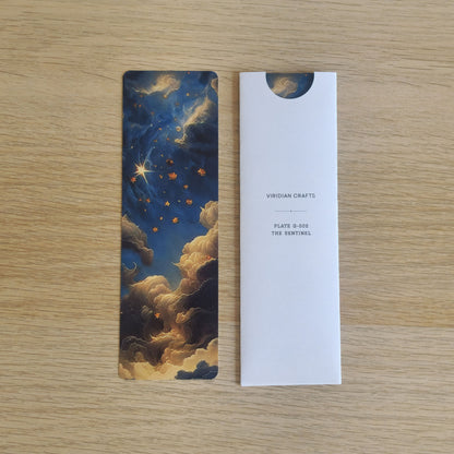 A rectangular bookmark with an illustrated night sky and clouds in dark blue, gold, and cream, placed beside a bookmark enclosed in a white protective paper sleeve printed with the brand name 'VIRIDIAN CRAFTS' and product text 'PLATE 8-602 THE SENTINEL' on a light wood surface.