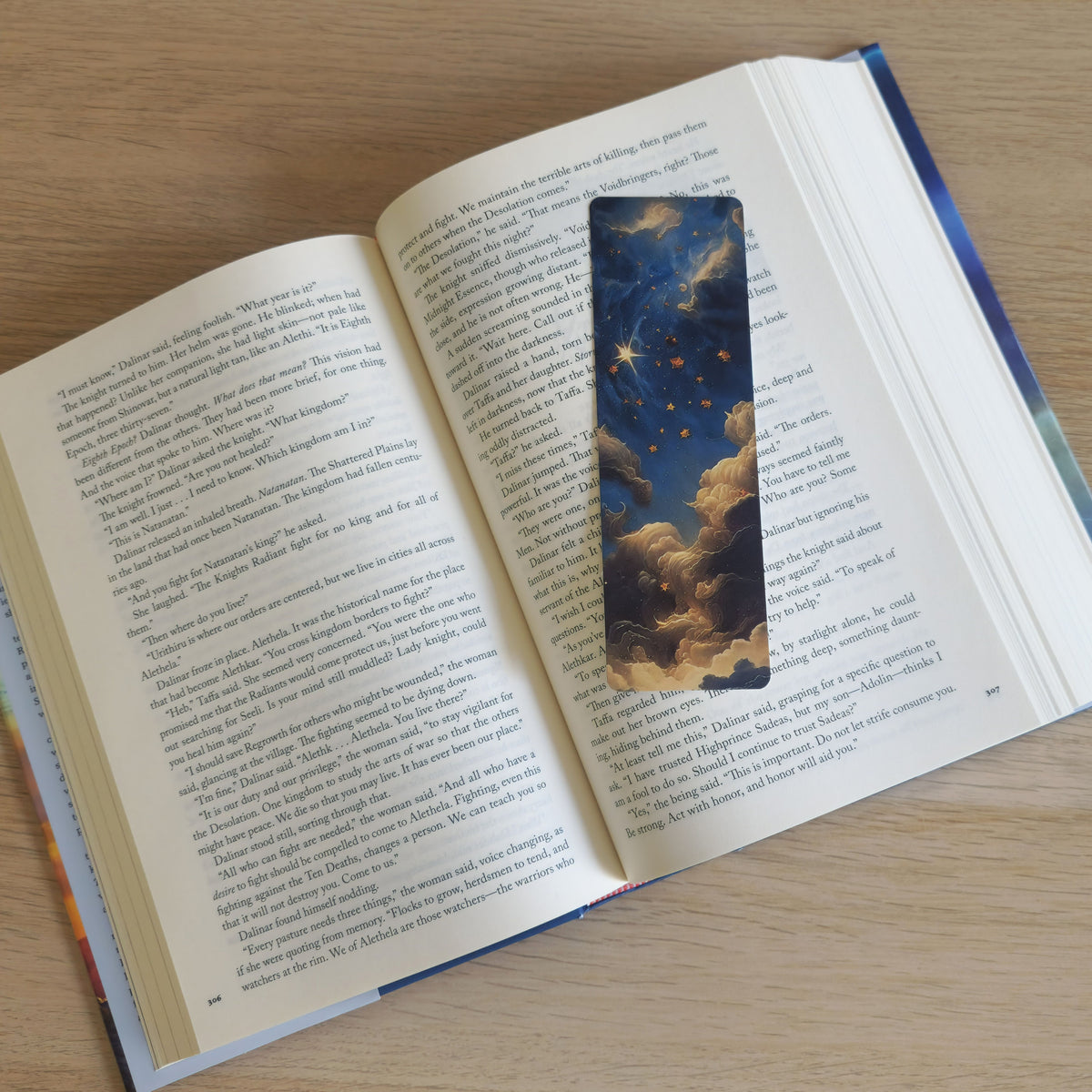 An open book on a light wood surface with a rectangular bookmark inserted between the pages. The bookmark depicts a starry night sky with clouds in blue, gold, and cream tones.