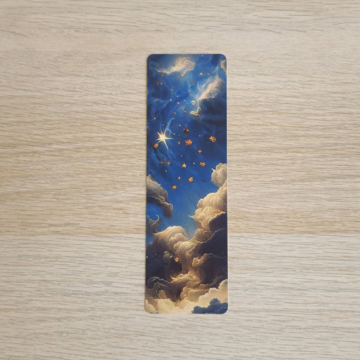 A slim rectangular book bookmark featuring a celestial night sky design with blue clouds, golden stars, and a bright shooting star, displayed on a light wood grain surface.
