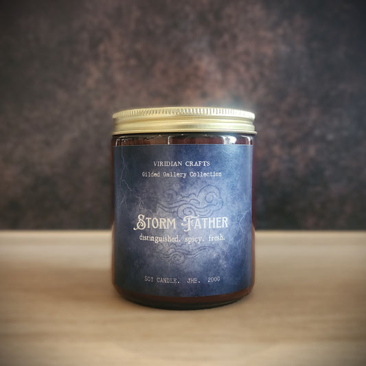 A glass jar candle with a gold metal lid and a dark blue textured label reading 'Storm Father' by Viridian Crafts, set on a light wooden surface against a dark textured background.