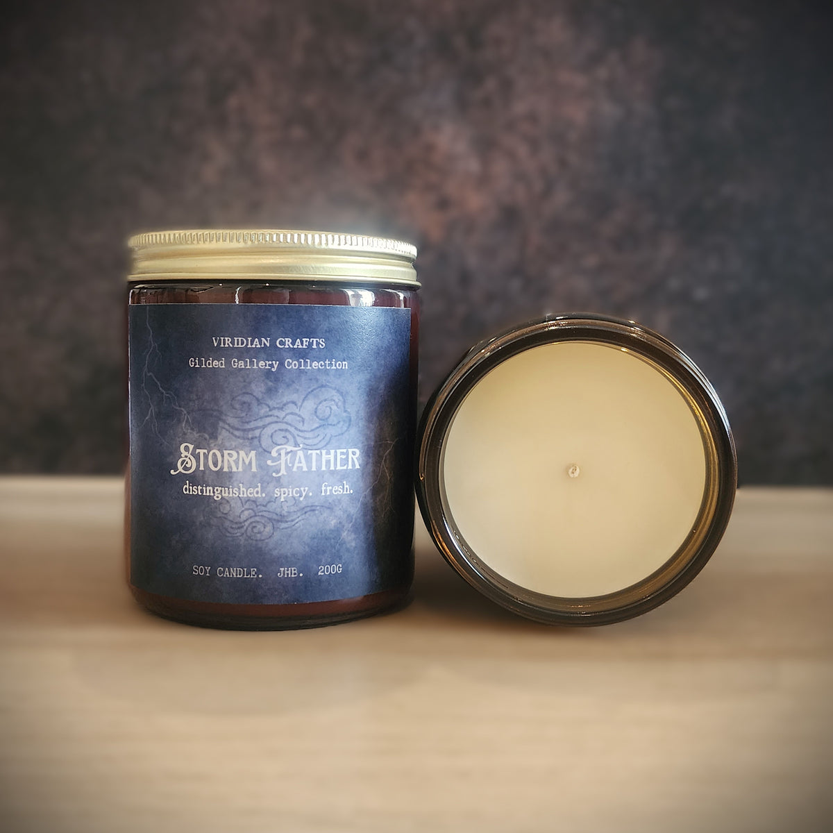 A soy candle in a dark amber glass jar with a blue label reading 'Storm Father' and 'Viridian Crafts Gilded Gallery Collection,' shown next to its open lid on a light wood surface against a dark textured background.