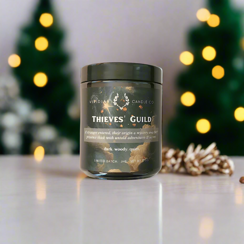 A Thieves' Guild candle inspired by Discworld and The Gentleman BAstard Series, handcrafted by Viridian Candle Co, placed on a table with Christmas lights and decorations in the background.