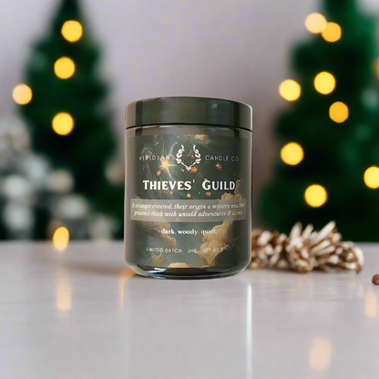 A Thieves' Guild candle inspired by Discworld and The Gentleman BAstard Series, handcrafted by Viridian Candle Co, placed on a table with Christmas lights and decorations in the background.