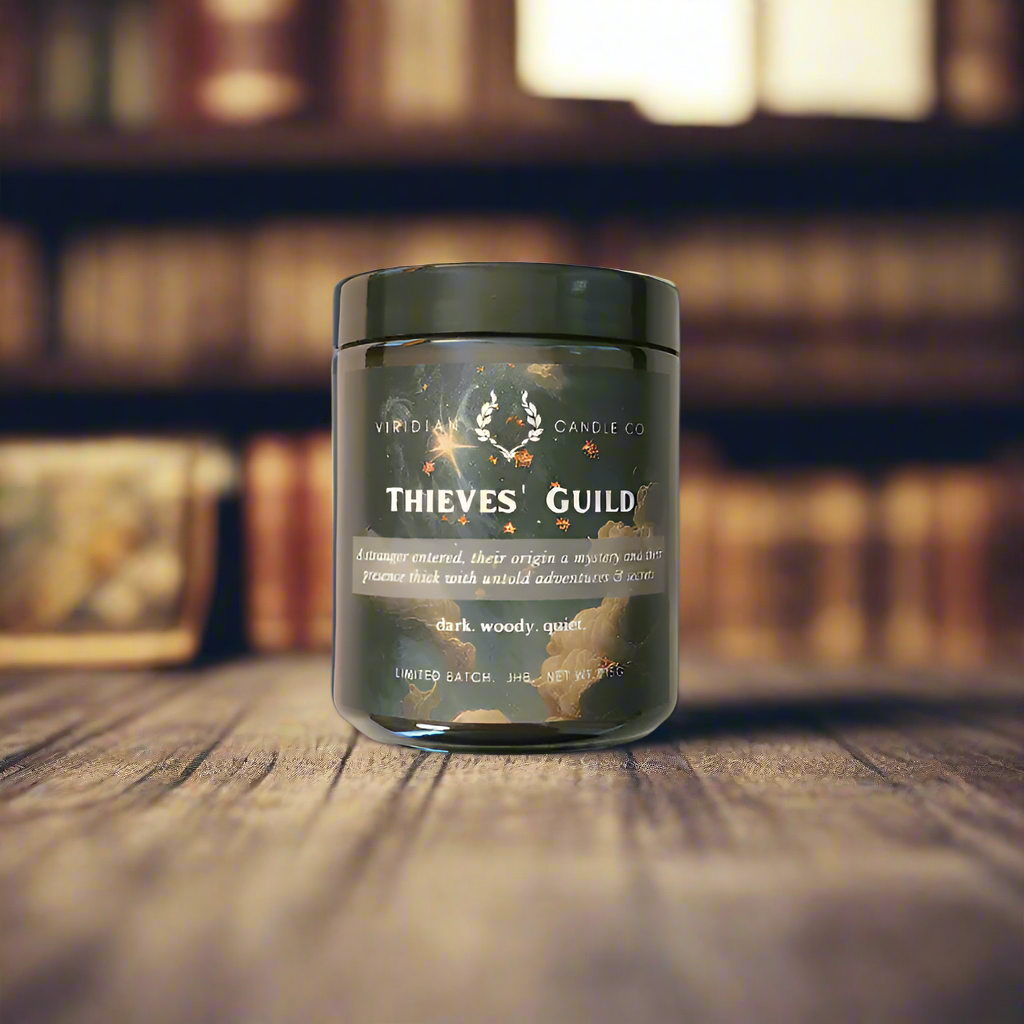 A Thieves' Guild candle, handcrafted by Viridian candle Co. on a wooden table with a bookcase in the background. Inspired by fantasy.