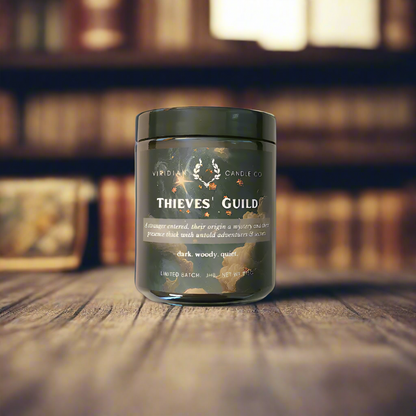 A Thieves' Guild candle, handcrafted by Viridian candle Co. on a wooden table with a bookcase in the background. Inspired by fantasy.